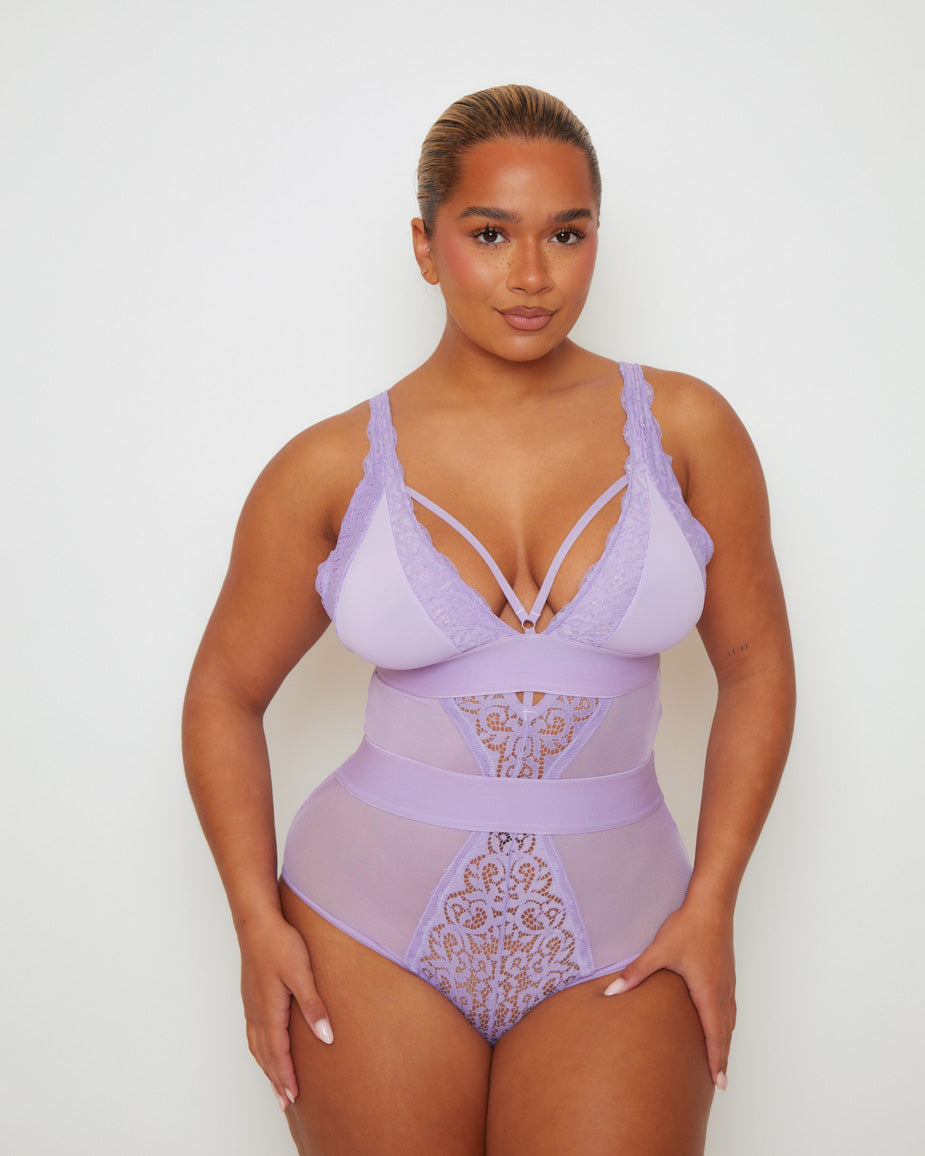 Model wearing the Alicia Fuller Bust Curvy Girl Bodysuit in Soft Lavender, featuring a plunging neckline with decorative strap detailing, floral lace cups, sheer mesh panels, and a high-waisted silhouette designed to flatter and support fuller busts.