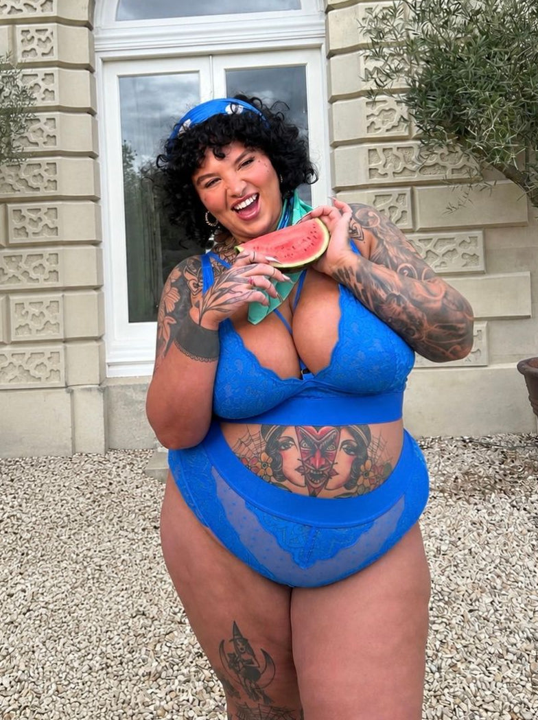 Model wearing the Hallie Bralette and Brief Set in Dazzling Blue from Tutti Rouge, posing outdoors while holding a slice of watermelon. The set features a supportive lace bralette with sheer mesh detailing and matching high-waisted briefs, styled with a blue headscarf.